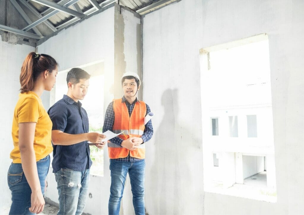 Three people, including a construction worker in safety gear, discussing plans inside an unfinished commercial building, addressing deferred maintenance issues.