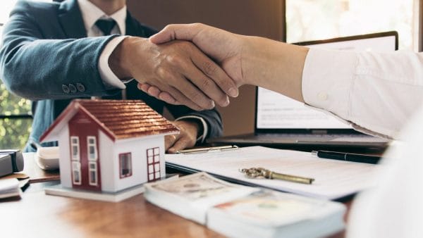 Two individuals shake hands over a desk with a model house, keys, and financial documents, indicating a real estate transaction, possibly related to a blog discussing the implications of a NAR lawsuit on commission practices in the real estate industry.