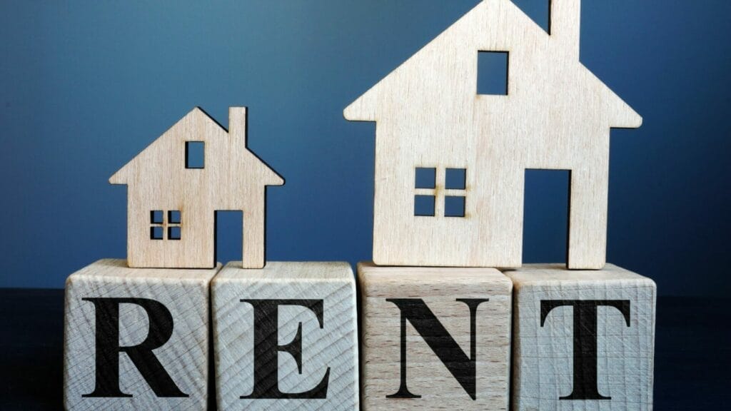 Wooden blocks spelling out 'RENT' with two wooden house shapes on top, against a blue background, symbolizing rental income as a key aspect of commercial property management fee analysis.