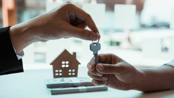 One person hands over a house key to another, symbolizing the transfer of property ownership, with a model house and financial documents in the background, an image potentially used in discussions about the NAR lawsuit's impact on home buyers and sellers.