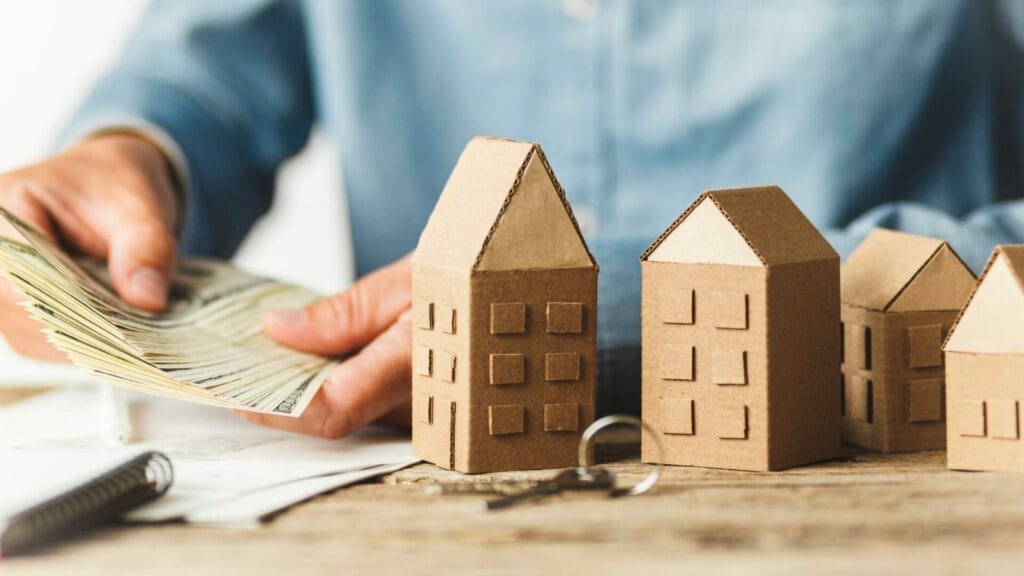 A person counts a stack of money next to a lineup of cardboard house models, symbolizing investment and financial planning in real estate, potentially linked to a discussion on maximizing operational profit, commonly referred to as net operating income or NOI, in property management.