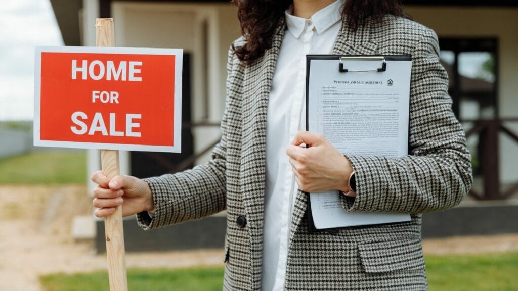 A real estate agent holds a "HOME FOR SALE" sign in one hand and a clipboard with a purchase and sale agreement in the other, indicative of the real estate market activity and possibly relating to a blog post about the impact of a NAR lawsuit on real estate practices.