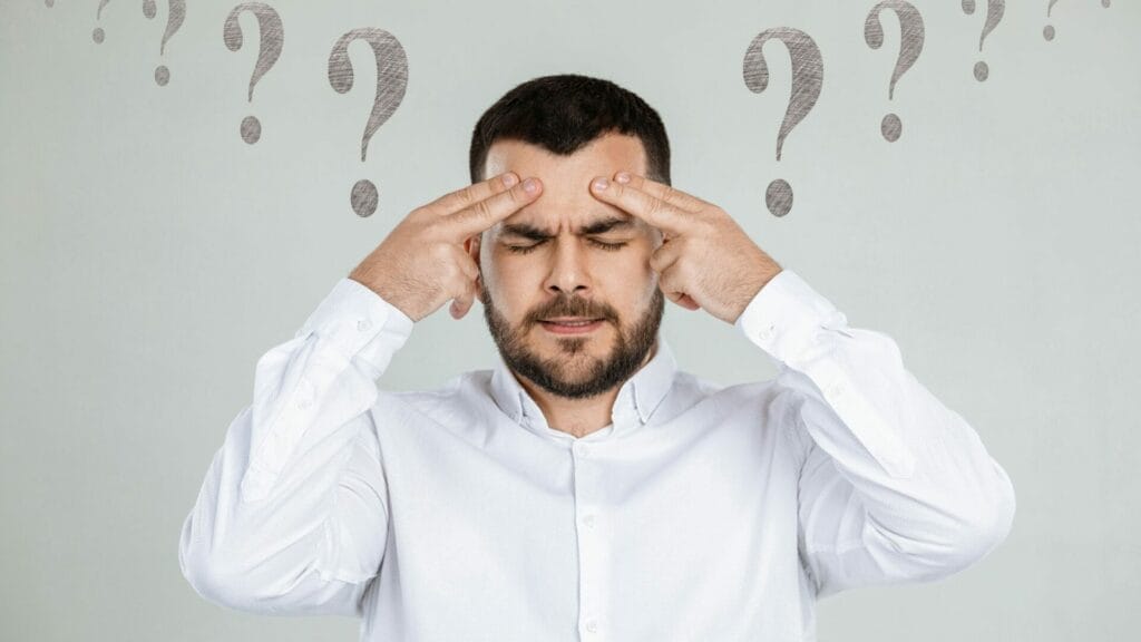 A perplexed man in a white shirt, with a furrowed brow and hands on his temples, surrounded by question marks, symbolizing the common confusion addressed in "Frequently Asked Questions on How to Value Commercial Property".