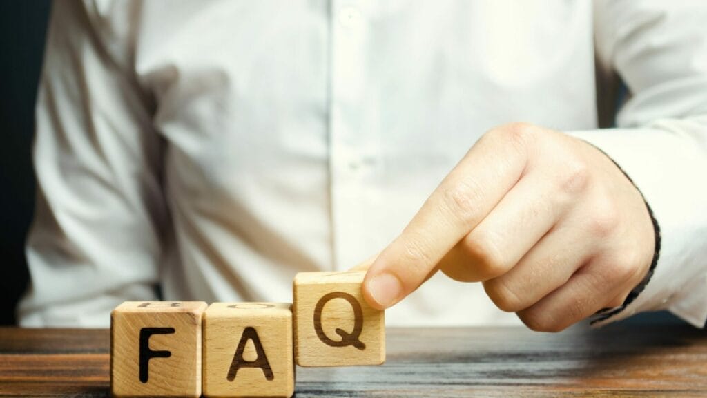 A person's hand arranging wooden blocks with letters to spell out "FAQ", representing frequently asked questions on Modified Gross Lease, set against a dark background with a white shirt visible, emphasizing a professional context.
