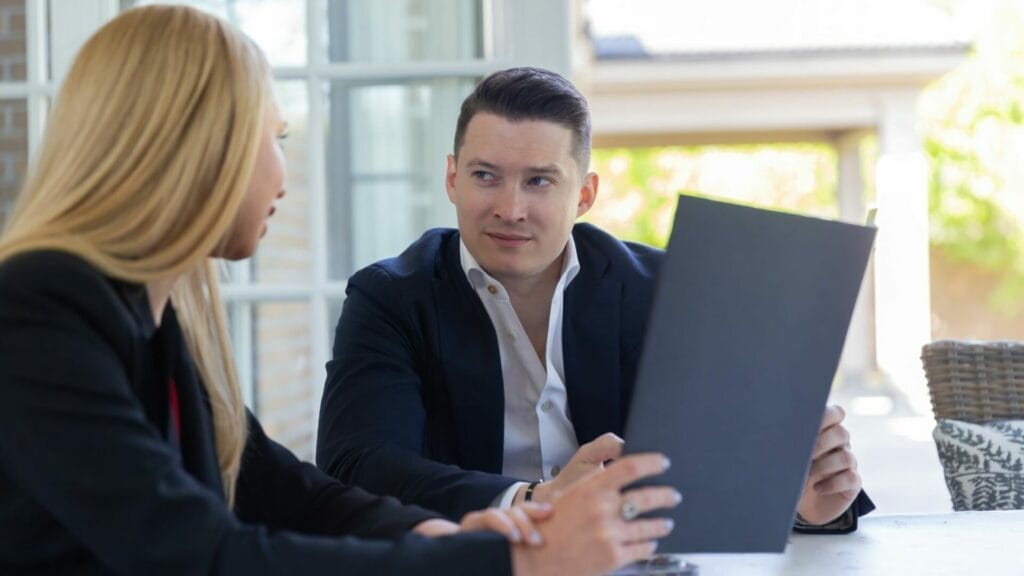 Two professionals in a serious discussion, with one holding and reviewing a document which could represent the key components of a Modified Gross Lease. They are seated at a table in a room with ample natural light and a casual, modern decor.