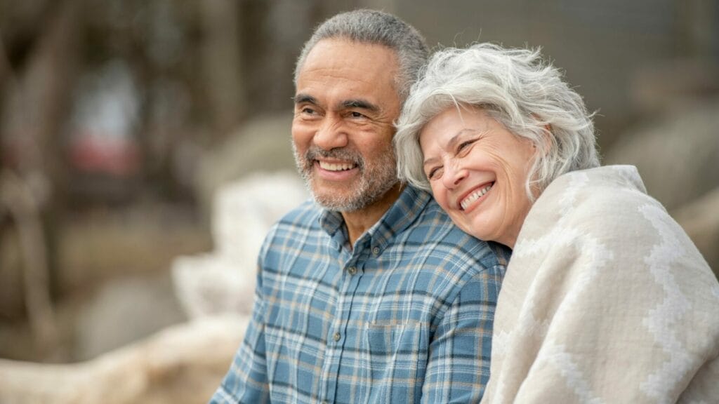 A joyful senior couple, wrapped in a cozy blanket, sitting outdoors and smiling broadly, embodying the happiness of successfully selling their investment property before retirement.