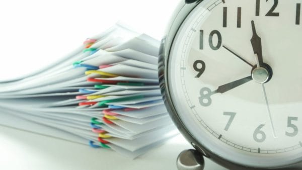A stack of paperwork with multi-colored paper clips is blurred in the background, symbolizing a pile of 1031 exchange forms, while a black vintage clock in the foreground indicates the time, perhaps referencing the critical deadlines associated with the 1031 exchange process. The time on the clock is close to nine, possibly denoting the urgency and time-sensitive nature of filing the necessary tax documentation.