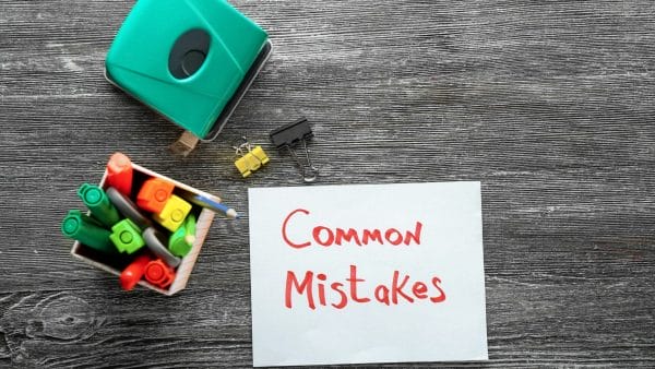 A piece of paper with "Common Mistakes" written in red marker rests on a textured wood surface, accompanied by a green tape dispenser, colorful markers in a holder, a pencil, and two paper clips. The image could represent a checklist or a warning about frequent errors to avoid when filling out a 1031 like-kind exchange form, emphasizing the importance of attention to detail in financial documentation.