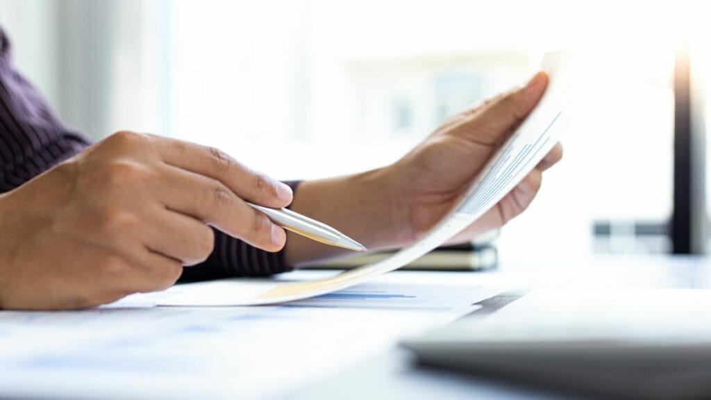 A person's hands are shown reviewing documents, potentially related to a 1031 tax-deferred exchange form. One hand holds the papers while the other uses a pen to point at or track information crucial to the 1031 property exchange process. The focus on the hands and documents suggests thorough scrutiny and the importance of accuracy in completing the tax forms for a real estate exchange. The setting is a bright office, implying a professional environment.